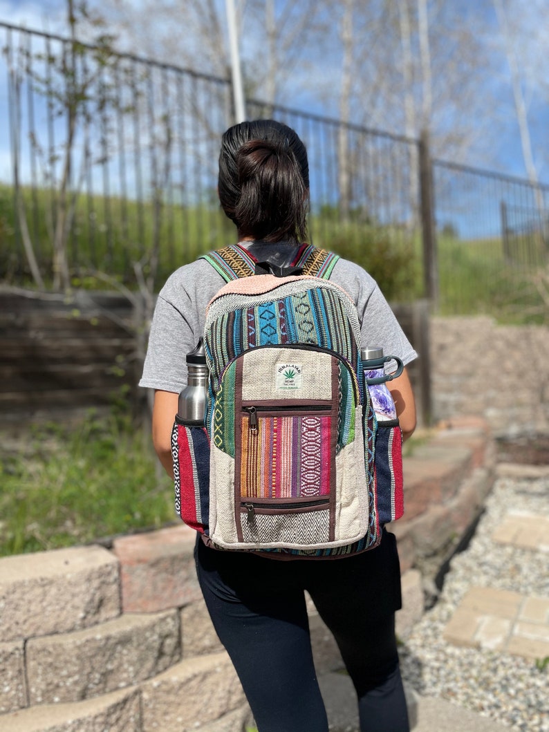 May include: A colorful backpack with a hemp leaf logo, featuring a mix of woven patterns in blue, red, and brown. It has side pockets holding a silver water bottle and a purple-wrapped item. The backpack is worn by a person in a gray shirt and black pants.