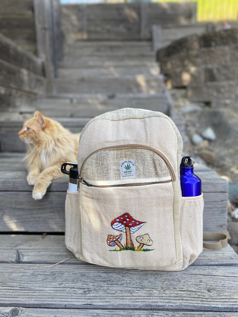 May include: A beige hemp backpack with a zippered pocket and a mushroom embroidery design. The backpack has the text "Himalayan Hemp" and "Made in Nepal" on a brown tag.