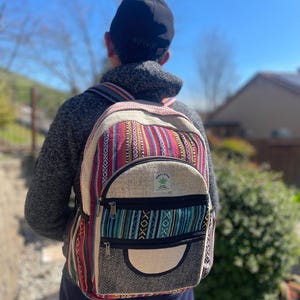 May include: A colorful backpack with a striped pattern in red, pink, and blue. The backpack has a front pocket with a black zipper and a hemp leaf logo. The bag is made of a woven material and has adjustable straps.
