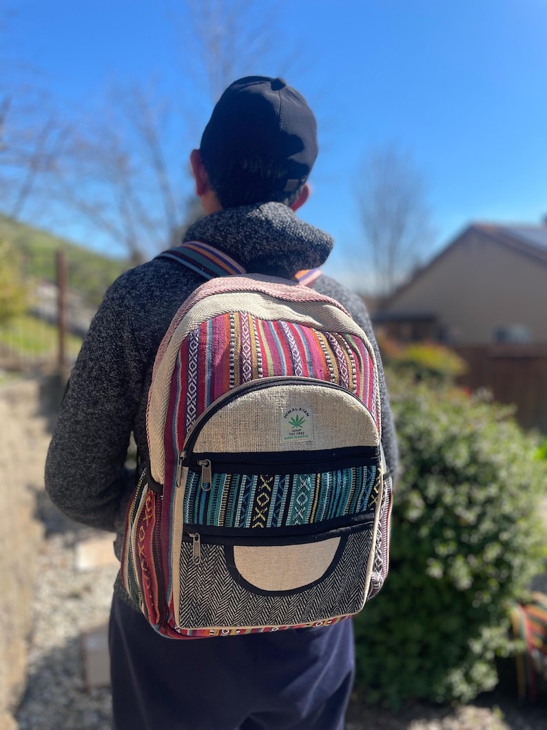May include: A colorful backpack with a striped pattern in red, pink, and blue. The backpack has a front pocket with a black zipper and a hemp leaf logo. The bag is made of a woven material and has adjustable straps.