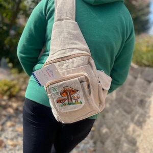 May include: A beige hemp sling bag with a zipped pocket and a patch featuring a mushroom design. The patch has the text "Himalayan Hemp" and "100% Hemp" on it.