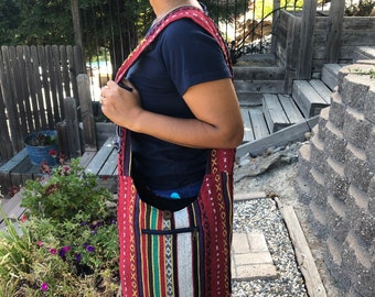 Può includere: Una borsa a tracolla in tessuto a righe rosse, nere, bianche e gialle con una tasca con cerniera. La borsa ha una tracolla lunga e si porta sulla spalla.
