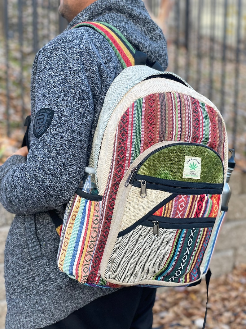 May include: A multi-coloured hemp backpack with a green patch that says "Himalayan Hemp" and a marijuana leaf logo. The backpack has a zipped pocket on the front and two side pockets.