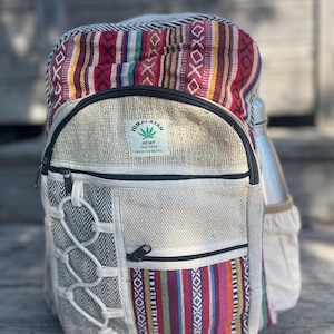 May include: Himalayan hemp backpack with a colorful striped top section and a neutral-toned body. Features a side pocket for a water bottle and a front pocket with a zipper. The backpack has a label that reads "Himalayan Hemp THC Free Made in Nepal."