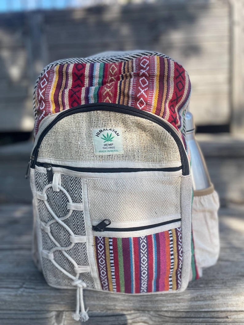 May include: Himalayan hemp backpack with a colorful striped top section and a neutral-toned body. Features a side pocket for a water bottle and a front pocket with a zipper. The backpack has a label that reads "Himalayan Hemp THC Free Made in Nepal."