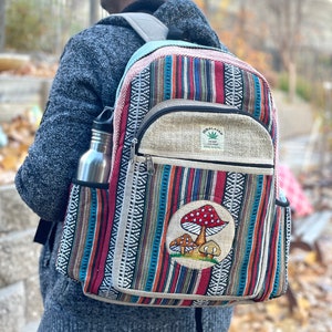 K&ouml;nnte beinhalten: Ein mehrfarbiger, gestreifter Hanfrucksack mit einer gro&szlig;en Fronttasche, die mit einem Pilzmotiv und dem Text "Hemp Incense" in einem gr&uuml;nen Blattdesign verziert ist. Der Rucksack hat eine Seitentasche f&uuml;r eine Wasserflasche.