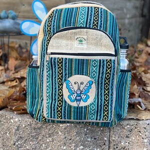 May include: A blue and white striped hemp backpack with a butterfly embroidered on the front. The backpack has a zippered pocket and a small label that reads "Himalayan Hemp Eco-Friendly".