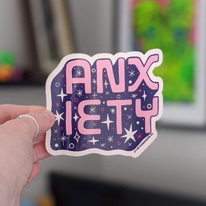 May include: A pink and blue sticker with the words "ANXIETY" in pink letters on a dark blue background. The sticker is decorated with white stars and other small shapes.