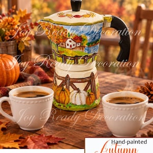 May include: A hand-painted stovetop espresso maker featuring an autumn landscape with a house, trees, and pumpkins on the base. The cream-coloured pot has a black handle and lid knob, accompanied by two white ceramic mugs filled with dark coffee.