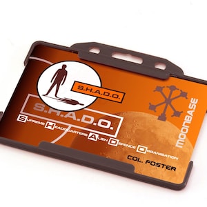 May include: A black plastic ID card holder with a clear plastic insert. The card inside has an orange background with the text "S.H.A.D.O. SUPREME HEADQUARTERS ALIEN DEFENCE ORGANISATION MOONBASE COL. FOSTER" and a stylised image of a person and a star.