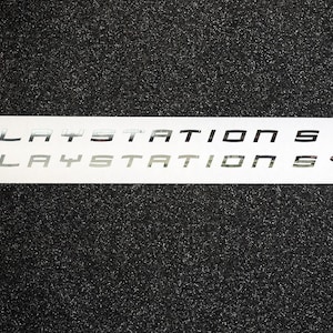 May include: A white sticker with the text "PLAYSTATION 5" in silver lettering, repeated twice. The PlayStation logo is in the top right corner of the sticker.