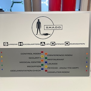 Può includere: Un cartello bianco con una silhouette nera di una persona in piedi su una piattaforma. Il cartello recita "S.H.A.D.O." e "SUPREME HEADQUARTERS ALIEN DEFENCE ORGANISATION". Sotto il testo ci sono cinque stanze numerate: Sala di controllo, Sicurezza, Centro medico, Ricevimento e Controllo della documentazione.