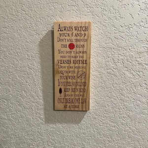 May include: Wooden sign with engraved text in a dark brown color. The sign reads "ALWAYS WATCH YOUR 6 AND 9" and other humorous advice. The sign is rectangular and mounted on a textured wall.