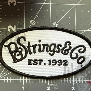 Pu&ograve; includere: Toppa ovale con sfondo grigio chiaro e bordo nero. La toppa presenta il testo "Strings & Co" in un font stilizzato, con "EST. 1992" sotto. La toppa &egrave; probabilmente per abbigliamento o accessori.