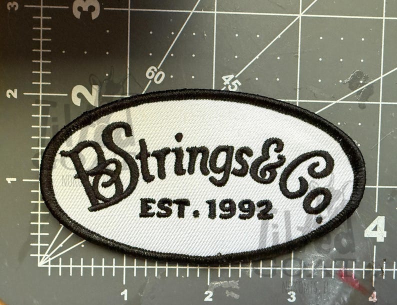 Pu&ograve; includere: Toppa ovale con sfondo grigio chiaro e bordo nero. La toppa presenta il testo "Strings & Co" in un font stilizzato, con "EST. 1992" sotto. La toppa &egrave; probabilmente per abbigliamento o accessori.