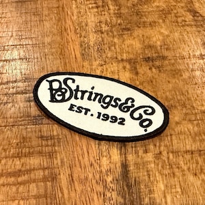 May include: An oval patch with the text "B Strings & Co. EST. 1992" in black script on a white background. The patch has a black border and is set against a wooden surface.