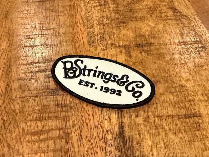 Pu&ograve; includere: Una toppa ovale con la scritta "B Strings & Co. EST. 1992" in nero su sfondo bianco. La toppa ha un bordo nero ed &egrave; posta su una superficie di legno.
