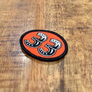May include: Oval patch with the number 33 in white, stylized with horseshoe details, on an orange background. The patch has a black border. The patch is on a wooden surface.