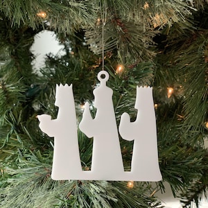 Puede incluir: Adorno de Navidad de silueta blanca que representa a los tres reyes magos, cada uno con una corona y sosteniendo un regalo. El adorno está colgado de un árbol de Navidad verde.