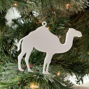 May include: White camel Christmas ornament hanging on a green Christmas tree. The ornament is made of a lightweight material and has a simple design.