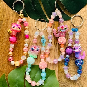 May include: Four colorful beaded keychains with charms. Each keychain features a different character, including a pink cat, a blue mermaid, a pink unicorn, and a purple dragon. The keychains are made with a variety of beads, including clear, iridescent, and glitter beads. The keychains are strung on a silver chain with a silver key ring.