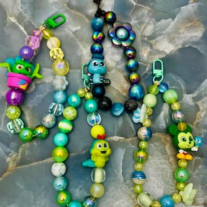 May include: Colorful beaded keychains with cartoon character charms. The keychains feature a variety of beads in shades of green, blue, yellow, and purple. Charms include a green alien, a blue character, a yellow caterpillar, and a Mickey Mouse-like character.