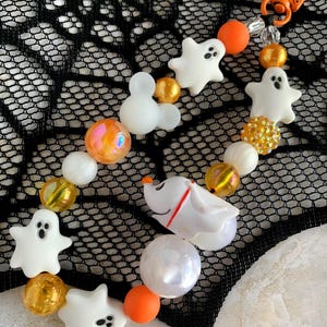 Może przedstawiać: Breloczek z koralików o tematyce Halloween z białymi zawieszkami duchów i postacią Zero, pomarańczowymi i złotymi koralikami oraz zapięciem na karabińczyk. Breloczek jest umieszczony na czarnym tle przypominającym pajęczynę.