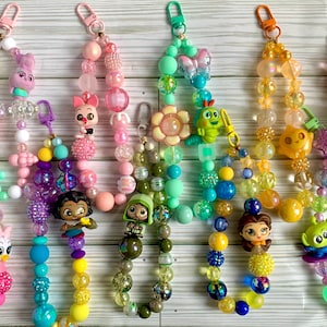 May include: A set of ten colorful beaded keychains with various cartoon character charms. The charms include a bunny, a flower, a bug, a princess, a mermaid, a spaceship, a dinosaur, a bear, a cat, and a dog.