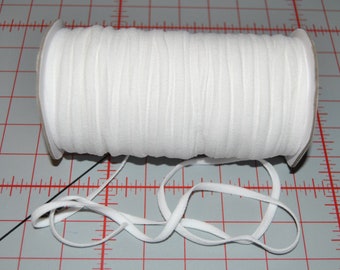 May include: A spool of white elastic cord, approximately 1/4 inch wide, is shown on a gray gridded surface. A length of the elastic cord is lying next to the spool.