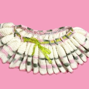 May include: A handmade, off-white crochet skirt with pink and green stripes. The skirt has a ruffled design and is tied with a bright green bow. The edges are finished with pink yarn, and the background is a solid pink.