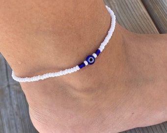 May include: White beaded anklet with a blue evil eye charm. The anklet is made of small white beads and features a central design of blue beads and an evil eye charm. The anklet is designed to be worn around the ankle.