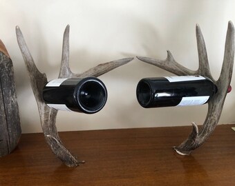 May include: A wine bottle holder made from two sets of deer antlers. The antlers are mounted on a wooden surface and are holding two bottles of wine.