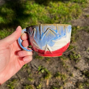 Op de afbeelding: Een keramische mok met een met de hand uitgesneden zigzagpatroon aan de buitenkant. Het glazuur verloopt van een beige rand naar een blauw middengedeelte en een effen rode basis. De mok heeft één gebogen handvat en een ronde komvorm.