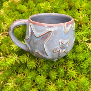 Könnte beinhalten: Eine handgefertigte Keramik-Tasse mit blau-roter Glasur und geprägten Sternenmustern. Die Tasse hat eine runde Form mit Henkel und steht auf einem Bett aus leuchtend grünem Laub. Ein einzigartiges Stück für Heißgetränke.