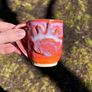 May include: A handmade ceramic mug with a vibrant orange and red glaze. The mug features a raised sun design with a smiling face and rays. The handle is curved and the mug is held by a hand.