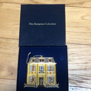 May include: A gold-colored house-shaped Christmas ornament in a blue velvet box. The box has the text "The Hampton Collection" on the lid.