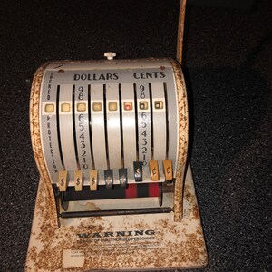 May include: A vintage white cash register with a black handle. The register has a display with the words "DOLLARS" and "CENTS" and a series of numbers. The register has a warning label that reads "BEWARE OF UNAUTHORIZED PERSONNEL".
