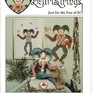 May include: A cross-stitch pattern featuring a jester with a red, white, and blue costume. The pattern is titled "HeartStrings Jest for the Fun of It!" and is part of "The Artists Collection" by Pat Thode.