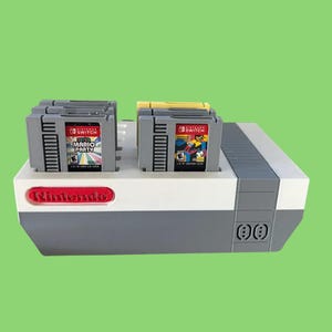 May include: A gray and white Nintendo Switch game cartridge holder with the Nintendo logo in red. Two gray game cartridges are inserted, one with "Mario Party" text. A yellow cartridge is also inserted.