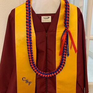 May include: A maroon graduation gown with a yellow stole and a blue and red lei. The lei has a blue ribbon with the year 2022 on it. The stole has the letters CSF embroidered on it.