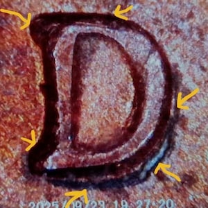 May include: Close-up of a copper-colored letter "D" with a textured surface. The letter has a dark outline and a lighter interior, with visible details of the metal's surface. The date and time are visible at the bottom.