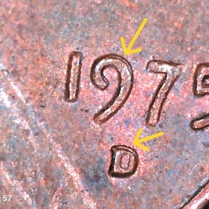 May include: A copper-colored metal coin surface featuring the embossed date 1975 and a D mint mark. Three thin yellow arrows point toward the number 9, the number 5, and the D mint mark to highlight specific details on the textured metal.