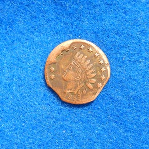 May include: A copper-colored coin with a profile of a Native American woman wearing a feathered headdress. The coin has a starburst design around the edge.