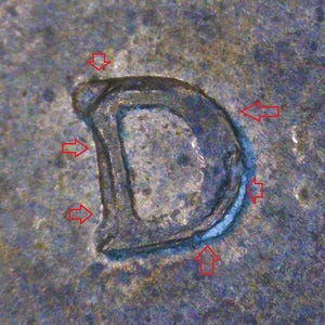 May include: A metallic surface featuring a stamped letter D with a visible double impression or doubling effect. Five red arrows point toward the edges of the letter, highlighting the secondary outline and the raised texture of the metal mark.