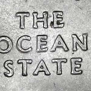 May include: Close-up of a silver coin with the words "THE OCEAN STATE" embossed on its surface. The text is in a bold, sans-serif font, and the coin has a textured background.