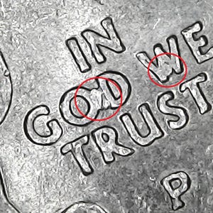 May include: Close-up of a silver coin with the words "IN GOD WE TRUST" and the letter "P" visible. The coin's surface has a textured, slightly worn appearance, with the lettering in a bold, clear font.