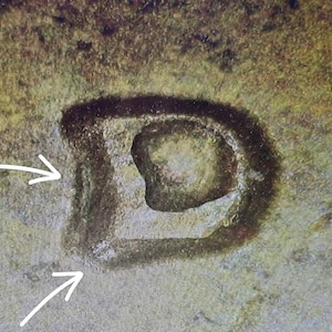 May include: A textured, dark brown, letter D-shaped mark embossed on a mottled, light brown surface. Two white, hand-drawn arrows point toward the left side of the letter, highlighting the edges of the dark, recessed, and textured character.