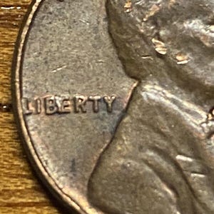 May include: Close-up of a copper penny with the word "LIBERTY" inscribed on the side. The image shows the profile of a person's face.
