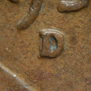 May include: Close-up of a bronze coin, showing the letter "D" in relief. The coin's surface has a textured, aged appearance with visible wear and tear. The details of the coin are highlighted by the lighting.