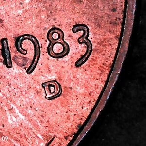 May include: A copper-colored coin surface featuring the embossed year 1983 and the mint mark D. The metal shows a textured, weathered patina with dark spots and a raised circular rim along the edge, captured in a high-magnification view.